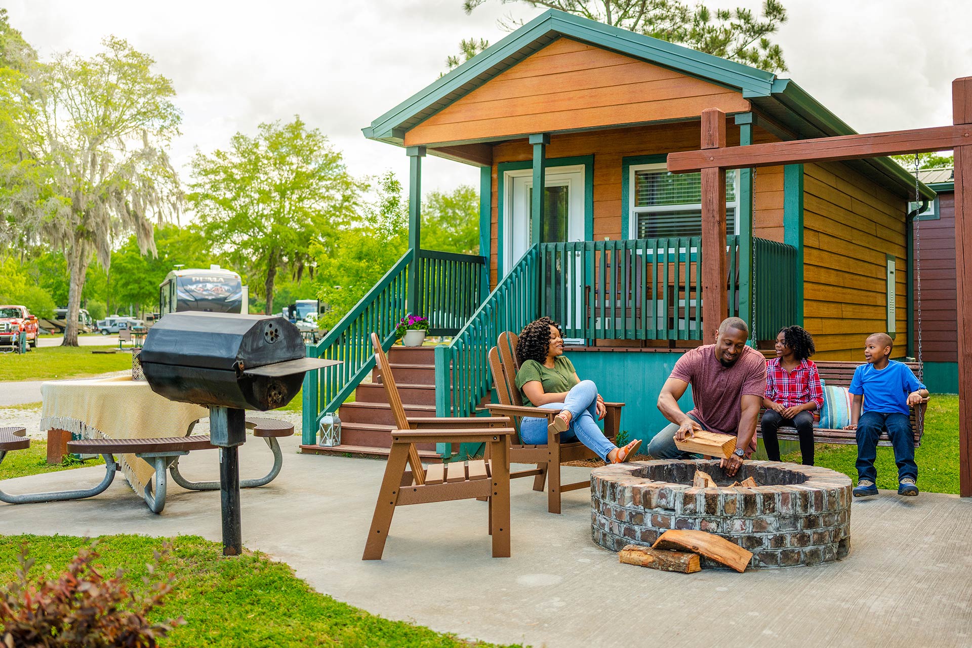 A family enjoys time together outside a cabin with a woman sitting on the porch, a man tending to a fire pit, and a woman and boy sitting on a bench. A picnic table with a grill and a lush green background with an RV are also visible.