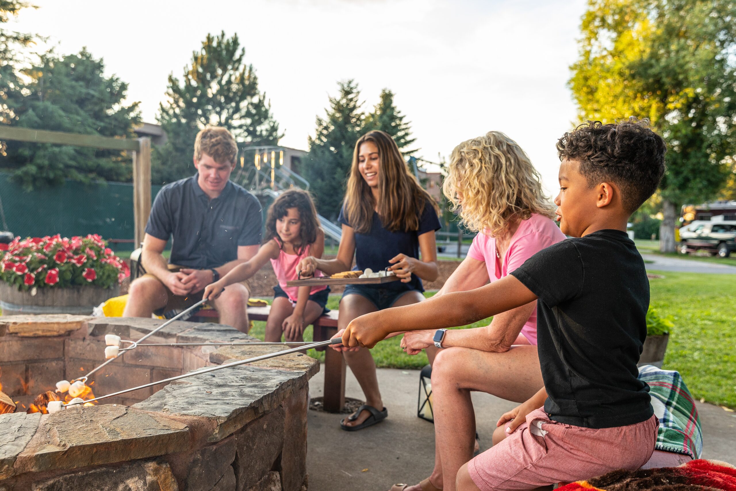 A group of people enjoying a marshmallow roasting activity around a fire pit in a backyard at dusk.
