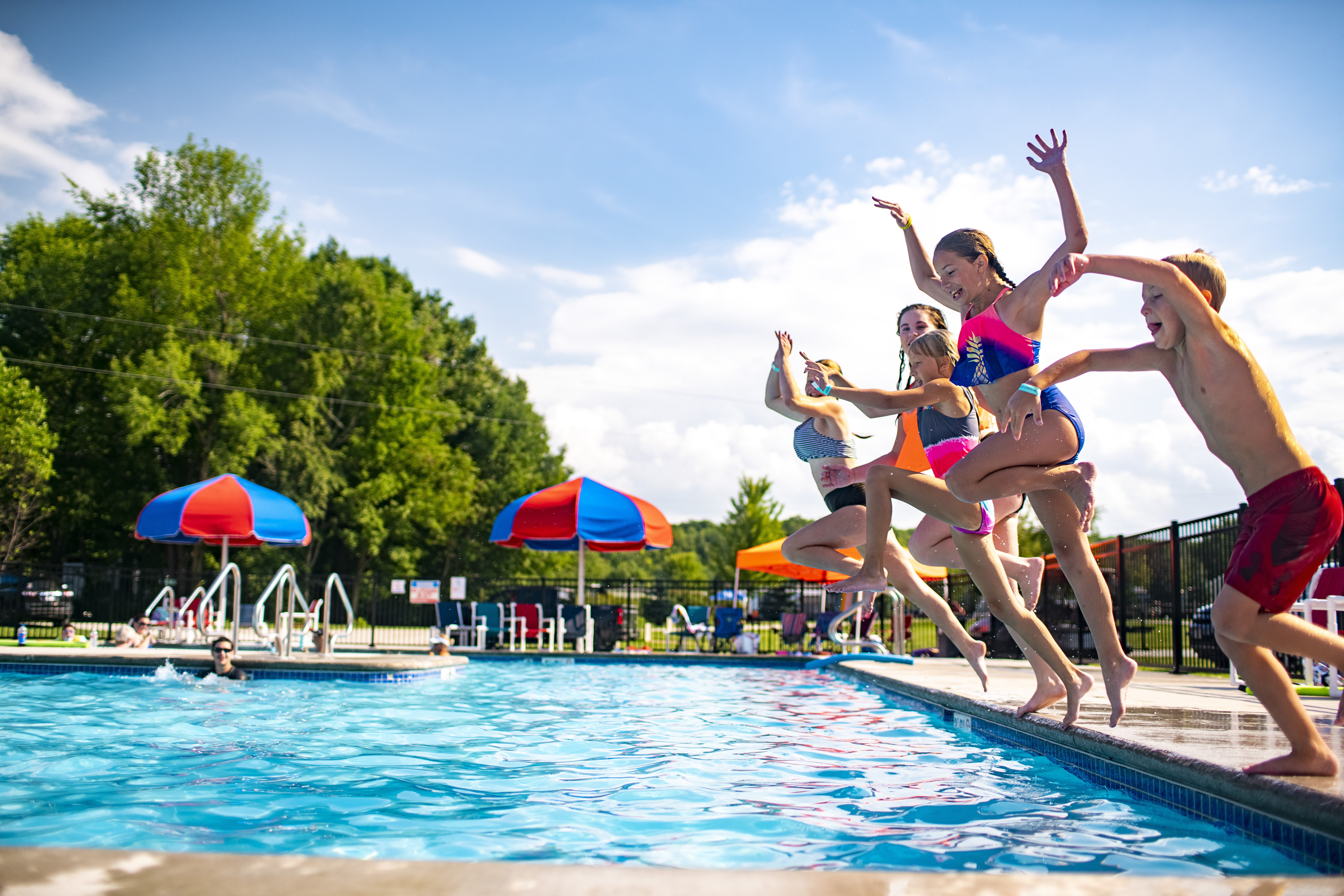 Children in swimsuits joyfully jumping into an outdoor swimming pool on a sunny day, with pool umbrellas, other swimmers, and a clear blue sky in the background.