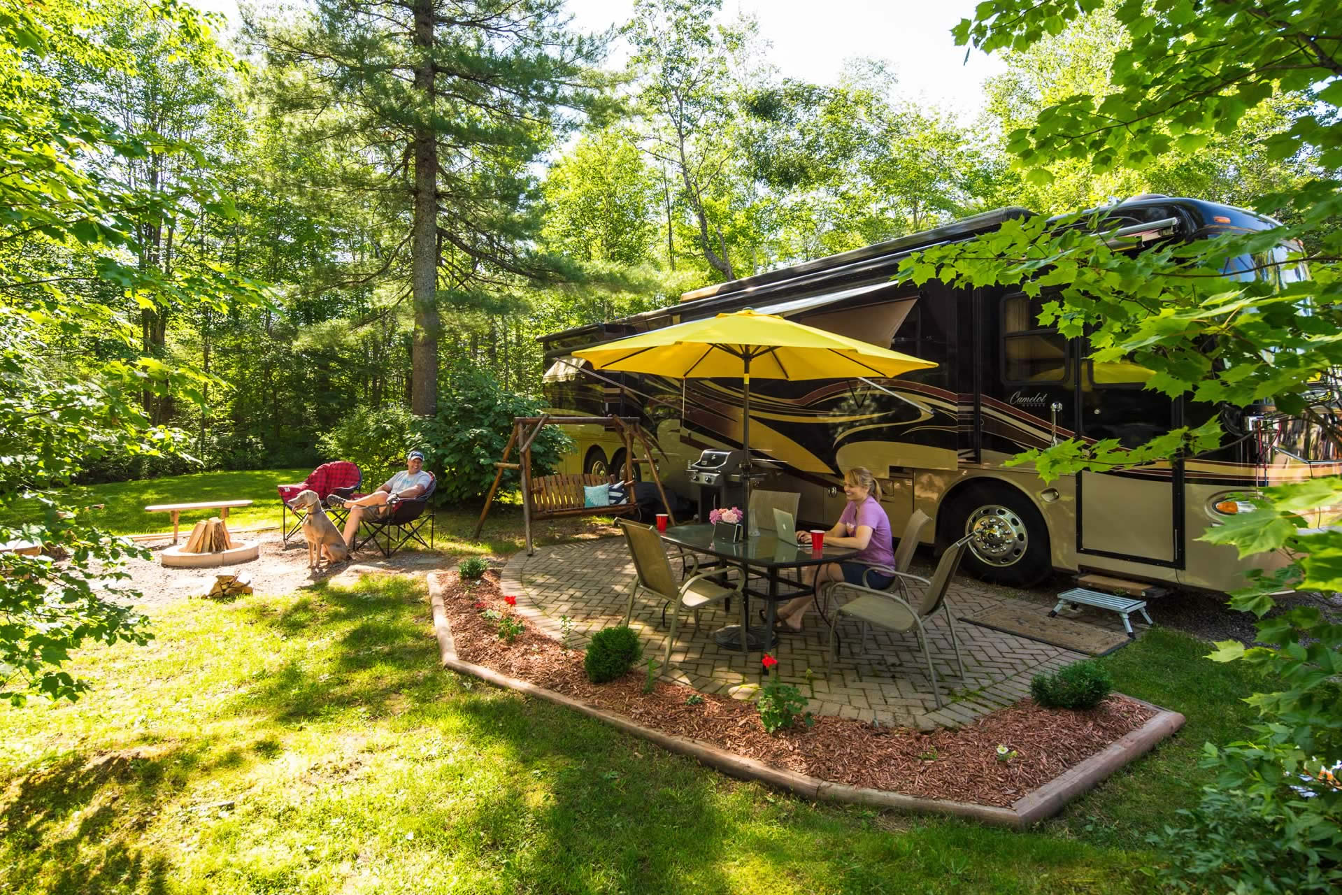 A peaceful campsite with a luxury RV and a yellow awning, where a woman works on a laptop at a table and a man sits with a dog by a fire pit in a wooded area.